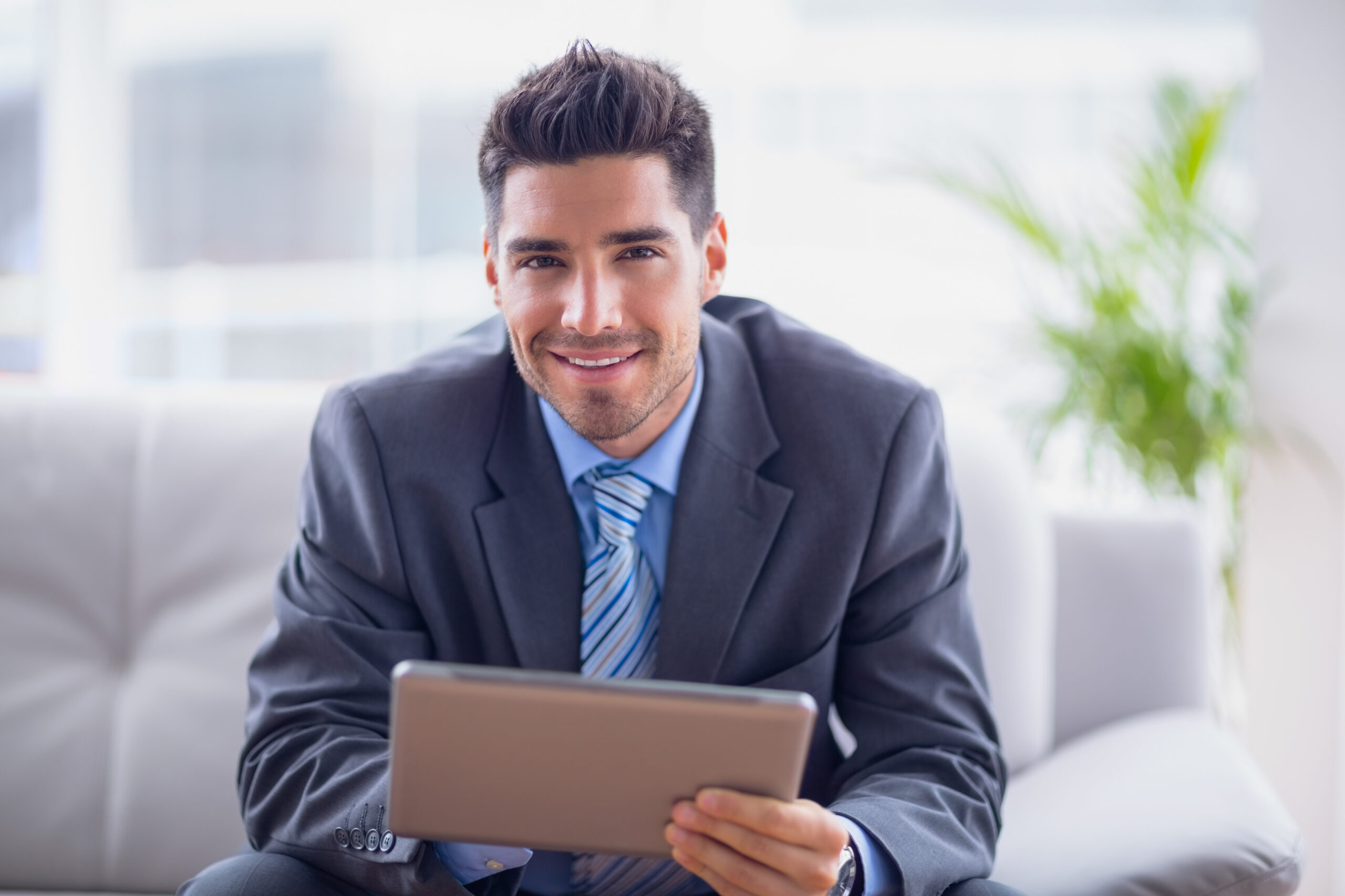 Businessman sitting on sofa using his tablet smiling at camera in the office